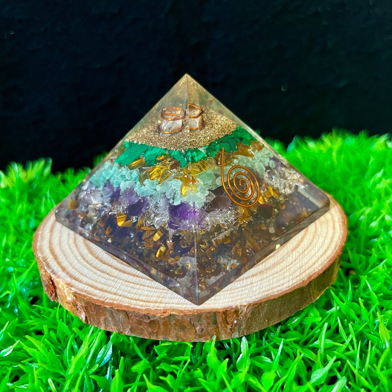 May include: A clear crystal pyramid with layers of green, purple, and gold crystals. The pyramid is sitting on a wooden base and is surrounded by green grass.