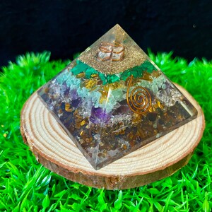 May include: A clear crystal pyramid with a copper wire spiral and various crystals embedded inside, including amethyst, tiger's eye, and malachite. The pyramid is resting on a wooden base.