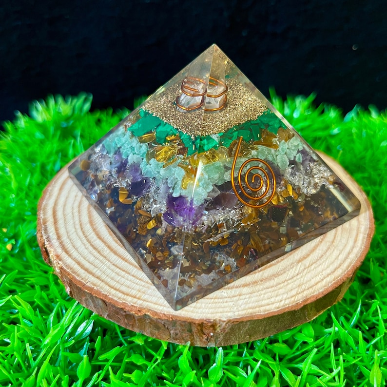 May include: A clear crystal pyramid with a copper wire spiral and various crystals embedded inside, including amethyst, tiger's eye, and malachite. The pyramid is resting on a wooden base.
