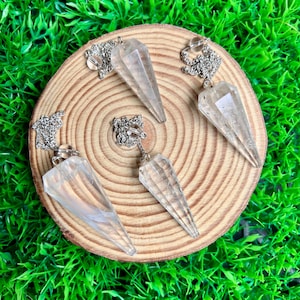 Op de afbeelding: Vijf heldere kwartskristallen pendels met zilverkleurige kettingen en decoratieve sluitingen worden getoond op een ronde houten schijf. De pendels zijn facetgeslepen en lopen taps toe naar een punt. De achtergrond is groen.