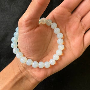 K&ouml;nnte beinhalten: Ein wei&szlig;es Opal-Armband mit durchscheinender, milchig wei&szlig;er Farbe. Das Armband besteht aus runden Perlen und ist auf einem Gummiband gef&auml;delt.