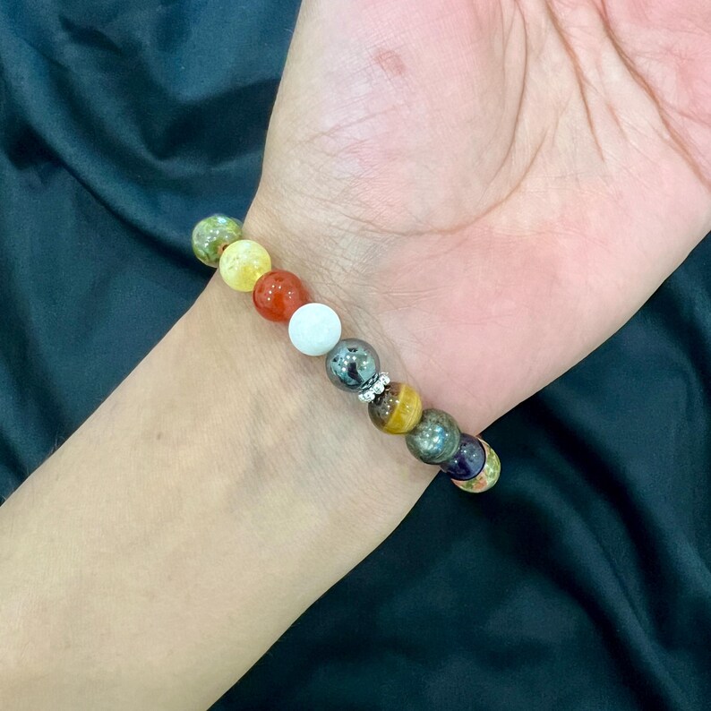 May include: A colorful beaded bracelet with a variety of stones, including green, yellow, red, white, black, brown, and purple. The bracelet is on a person's wrist.