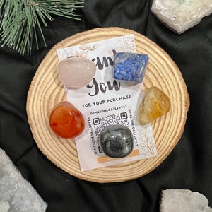 Puede incluir: Un conjunto de cinco piedras pulidas sobre una base de madera. Las piedras son cuarzo transparente, cornalina, lapislázuli, citrino y obsidiana negra. La base tiene un mensaje de agradecimiento y un código QR para GEMSTONEGALLERY733.