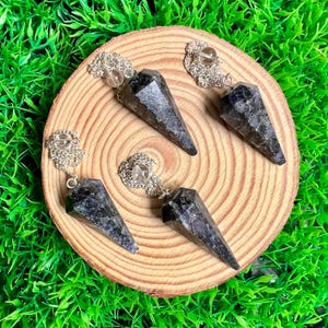 May include: Five dark gray crystal pendulums with silver chains and clear crystal accents are arranged on a round wooden slice. The pendulums are cone-shaped and have a textured surface. The background is bright green grass.