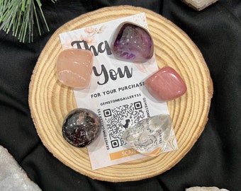 May include: A collection of polished gemstones in various colors, including pink, purple, and clear, arranged on a small wooden slice. A card with the words "Thank You" and a QR code is also present. The gemstones are likely for decorative or metaphysical purposes.