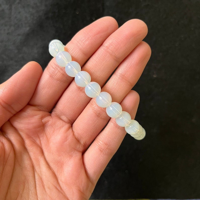 K&ouml;nnte beinhalten: Ein wei&szlig;es Opal-Perlenarmband mit einem dezenten irisierenden Schimmer. Das Armband besteht aus runden Perlen und ist auf einem Gummiband gef&auml;delt.