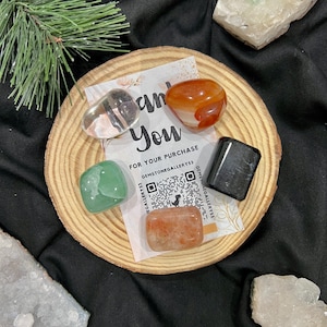 May include: A set of five tumbled stones on a wooden coaster. The stones are clear, green, orange, red, and black. The coaster has a thank you note with a QR code for Gemstone Gallery.