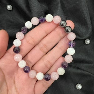 Könnte beinhalten: Ein Armband aus rosa, weißen und lila Perlen. Die Perlen sind rund und haben eine glatte, polierte Oberfläche. Das Armband ist auf einer durchsichtigen Gummikordel aufgefädelt.