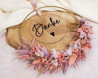 Puede incluir: Una corona de flores secas rosa y beige con un aro dorado y un letrero de acrílico transparente que dice "Danke" con un pequeño corazón.