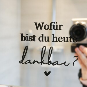 Könnte beinhalten: Eine Glasoberfläche mit schwarzem Text, der "Wofür bist du heute dankbar?" mit einem Herzsymbol lautet. Eine Kamera spiegelt sich im Glas. Der Text ist in einer Schriftart geschrieben.