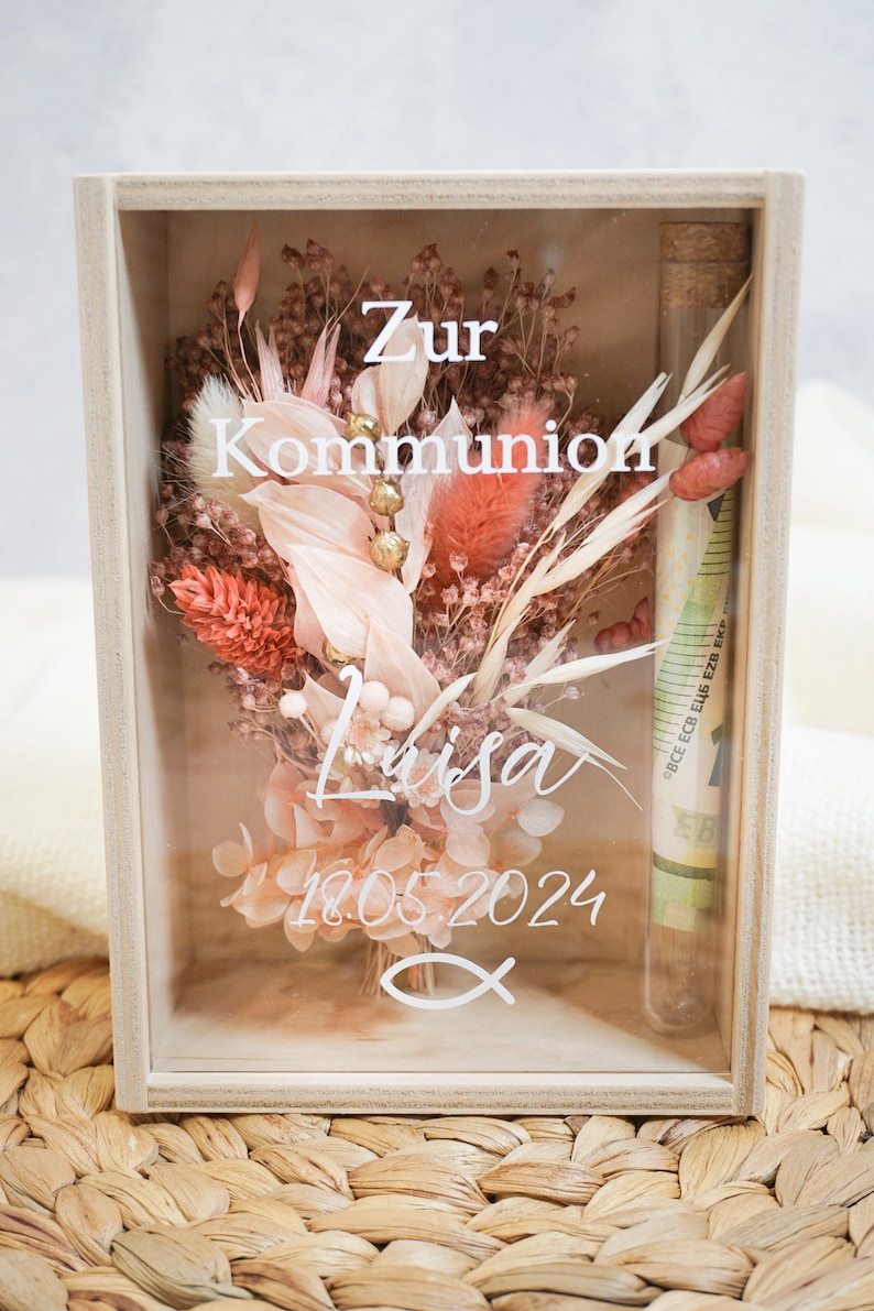 K&ouml;nnte beinhalten: Eine Holzkiste mit einer Glasfront, die getrocknete Blumen, ein kleines Glasr&ouml;hrchen mit Geld darin und den Text "Zur Kommunion Lisa 18.05.2024" auf dem Glas enth&auml;lt.
