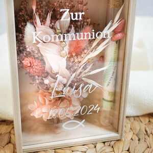 K&ouml;nnte beinhalten: Eine Holzkiste mit einer Glasfront, die getrocknete Blumen und ein kleines Glasr&ouml;hrchen mit einem eingerollten Geldschein enth&auml;lt. Die Kiste tr&auml;gt den Schriftzug "Zur Kommunion Lisa 18.05.2024".
