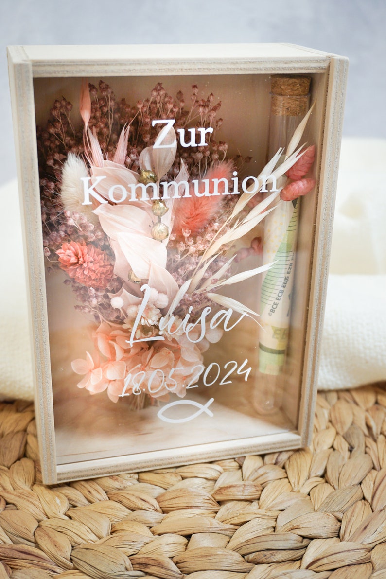 K&ouml;nnte beinhalten: Eine Holzkiste mit einer Glasfront, die getrocknete Blumen und ein kleines Glasr&ouml;hrchen mit einem eingerollten Geldschein enth&auml;lt. Die Kiste tr&auml;gt den Schriftzug "Zur Kommunion Lisa 18.05.2024".