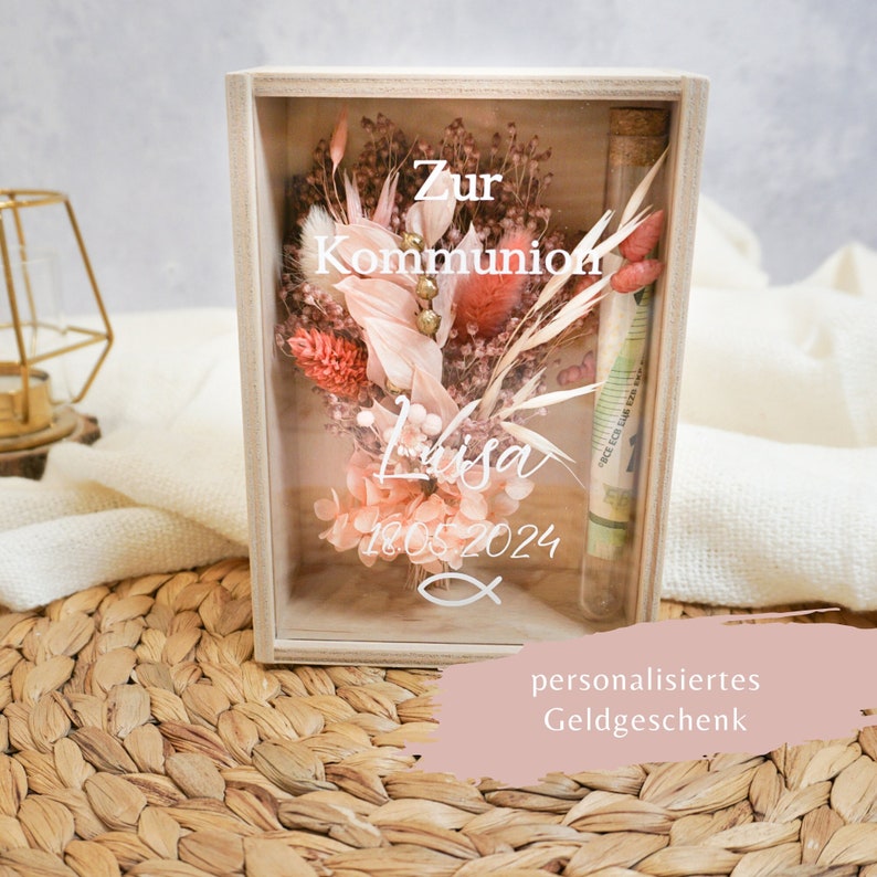 K&ouml;nnte beinhalten: Eine Holzkiste mit einer Glasfront, die getrocknete Blumen und ein kleines Glasr&ouml;hrchen mit Geld enth&auml;lt. Die Kiste tr&auml;gt den Text "Zur Kommunion Lisa 18.05.2024" und ein Fischsymbol. Der Text "personalisiertes Geldgeschenk" befindet sich unter der Kiste.