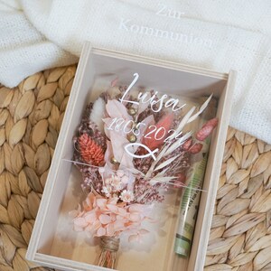 K&ouml;nnte beinhalten: Eine Holzkiste mit einem durchsichtigen Deckel, in der ein Trockenblumenarrangement liegt. Die Blumen sind in Rosat&ouml;nen, Pfirsich und Koralle gehalten. Die Kiste ist mit dem Text "Zur Kommunion Luisa 18.05.2024" beschriftet.