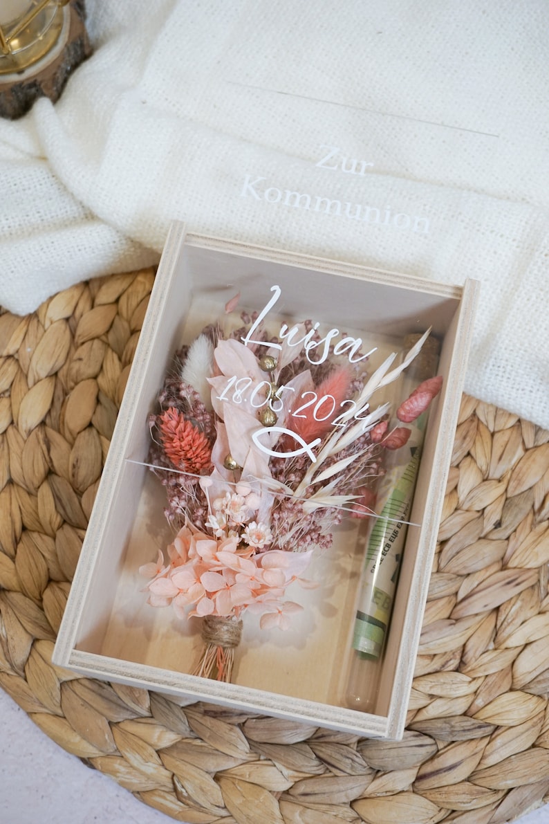 K&ouml;nnte beinhalten: Eine Holzkiste mit einem durchsichtigen Deckel, in der ein Trockenblumenarrangement liegt. Die Blumen sind in Rosat&ouml;nen, Pfirsich und Koralle gehalten. Die Kiste ist mit dem Text "Zur Kommunion Luisa 18.05.2024" beschriftet.