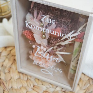 K&ouml;nnte beinhalten: Eine wei&szlig;e Holzkiste mit einer Glasfront, die getrocknete Blumen und eine kleine Glasflasche enth&auml;lt. Die Kiste tr&auml;gt den Text "Zur Kommunion Lisa 18.05.2024".