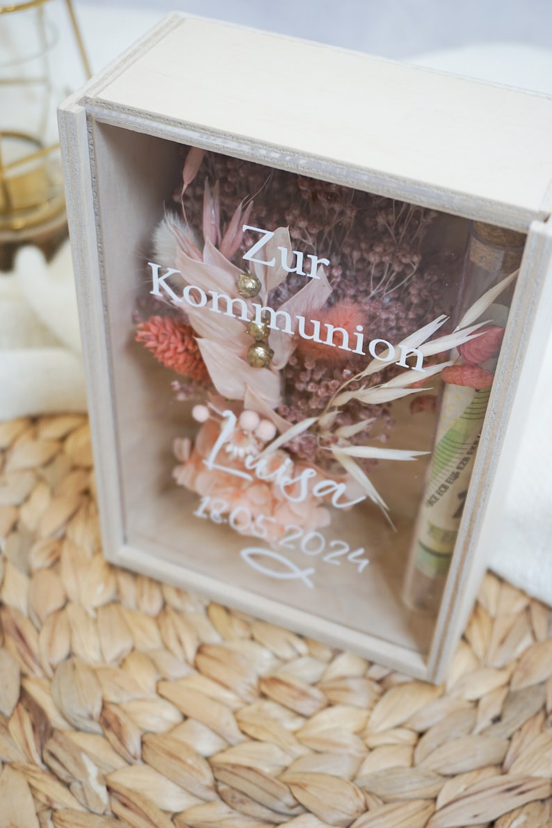 K&ouml;nnte beinhalten: Eine wei&szlig;e Holzkiste mit einer Glasfront, die getrocknete Blumen und eine kleine Glasflasche enth&auml;lt. Die Kiste tr&auml;gt den Text "Zur Kommunion Lisa 18.05.2024".