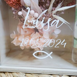 K&ouml;nnte beinhalten: Eine wei&szlig;e Holzkiste mit einer durchsichtigen Glasfront, die einen Trockenblumenstrau&szlig; enth&auml;lt. Die Blumen sind in Rosat&ouml;nen und Braun gehalten. Die Kiste ist mit dem Namen "Lisa" und dem Datum "18.05.2024" in wei&szlig; verziert. Ein wei&szlig;es Fischsymbol ist ebenfalls sichtbar.