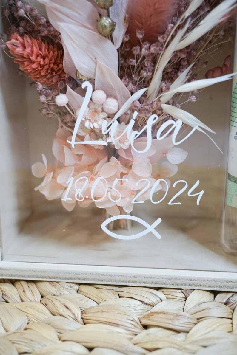 K&ouml;nnte beinhalten: Eine wei&szlig;e Holzkiste mit einer durchsichtigen Glasfront, die einen Trockenblumenstrau&szlig; enth&auml;lt. Die Blumen sind in Rosat&ouml;nen und Braun gehalten. Die Kiste ist mit dem Namen "Lisa" und dem Datum "18.05.2024" in wei&szlig; verziert. Ein wei&szlig;es Fischsymbol ist ebenfalls sichtbar.