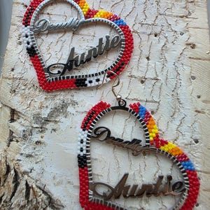 Peut inclure: Boucles d'oreilles en forme de cœur avec des bords perlés rouges, noirs, blancs, jaunes et bleus. Les boucles d'oreilles ont un cadre en métal argenté avec les mots "Deadly Auntie". Elles sont suspendues à des crochets argentés.