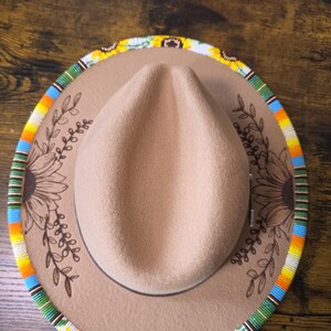 May include: A tan felt fedora hat featuring a wide brim adorned with a colourful beaded edge in yellow, green, white, and blue. The hat crown and brim are decorated with dark brown line art illustrations of sunflowers and leafy vine branches.