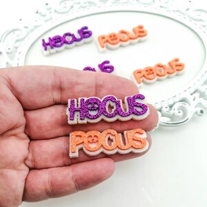 Pode incluir: Duas palavras cobertas de glitter, "Hocus" em roxo e "Pocus" em laranja, são seguradas em uma mão. As palavras são em relevo e têm um contorno branco.