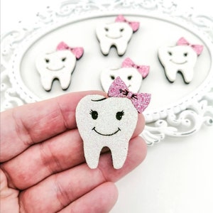 May include: A white glitter tooth with a pink bow, a smiling face, and eyelashes. The tooth is held in a hand. There are four other teeth in the background, also with pink bows.