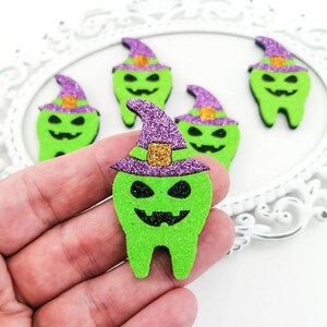 May include: Green foam Halloween teeth with witch hats. The teeth have black eyes and smiles. The witch hats are purple with gold glitter accents. The teeth are on a white decorative tray.