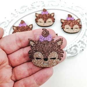 Op de afbeelding: Close-up van verschillende glitterende vossengezichtversieringen. De vossengezichten zijn bruin en bruin met gesloten ogen en een kleine glimlach. Elke vos heeft een paarse strik op zijn hoofd. Op een witte achtergrond.