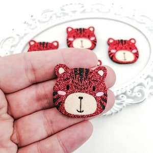 May include: Small red glitter tiger face embellishments with black stripes, pink cheeks, and a beige nose. The image shows several of these decorative tiger faces. They have a glittery texture.
