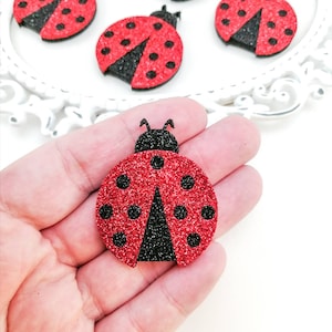 Puede incluir: Cinco formas de mariquita de purpurina roja y negra con puntos negros. Las mariquitas tienen un diámetro de aproximadamente 5 cm.