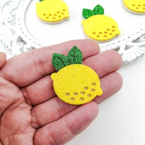 May include: Yellow glittery lemon-shaped embellishments with green glitter leaves. The lemons have small, evenly spaced holes along the bottom edge. The lemons are on a white surface.