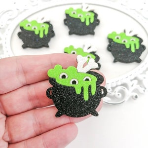 May include: Five black cauldron shaped Halloween decorations with green goo and googly eyes. The cauldrons are covered in black glitter and have a white bone on top.