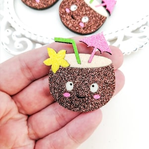 May include: A brown glitter coconut with a pink umbrella, green straw, and yellow flower. The coconut has a smiling face with pink cheeks and black eyes.