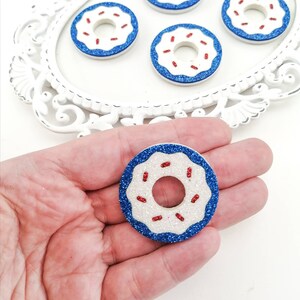 May include: Five blue and white glitter donuts with red sprinkles. The donuts are on a white tray with a decorative border. One donut is being held in a hand.