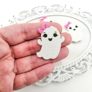 Op de afbeelding: Glitterende witte spookvormige versieringen met roze strikken. De geesten hebben zwarte ogen en lachende monden. De versieringen liggen op een wit decoratief dienblad.