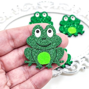 Könnte beinhalten: Grüne Glitzer-Frosch mit weißen Augen und grünem Bauch. Der Frosch sitzt auf einer Hand.