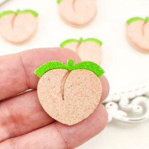 Puede incluir: Primer plano de adornos en forma de melocotón con una superficie brillante y texturizada. Cada melocotón tiene un detalle de hoja verde en la parte superior. Los melocottones son de color naranja claro y probablemente sean para manualidades o decoración.