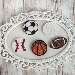 May include: Five white glitter sports ball ornaments with red, brown, and black details. The ornaments include a baseball, basketball, football, soccer ball, and volleyball.