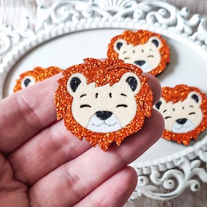 May include: Four orange glitter lion head cutouts with black eyes and white faces. The lion heads are on a white surface.