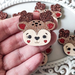 Può includere: Primo piano di diversi abbellimenti per il viso di renna glitterati. Ogni renna ha corna glitterate marroni, un viso color camoscio, guance rosse a forma di cuore e occhi neri. Le renne sono su un vassoio decorativo bianco.