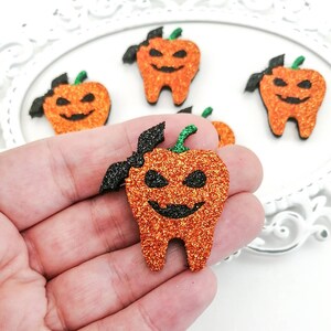 May include: Orange glittery Halloween-themed tooth-shaped embellishments. Each tooth features a jack-o'-lantern face with a black smile and eyes, a green stem, and a black bat wing. The teeth are displayed on a white background.