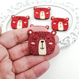Op de afbeelding: Rode glitter berenvormige versieringen met gesloten ogen, kleine oren en een crèmekleurige snuit. De berengezichten hebben roze wangen en een zwarte glimlach. Deze decoratieve stukken zijn ideaal voor knutsel- en scrapbookingprojecten.
