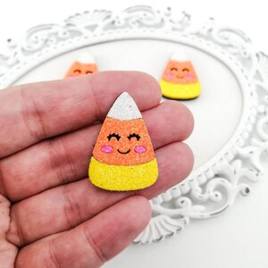 Może przedstawiać: Zestaw trzech ozdobnych kukurydzianych cukierków pokrytych brokatem z uśmiechającą się buzią. Cukierki kukurydziane są białe, pomarańczowe i żółte.