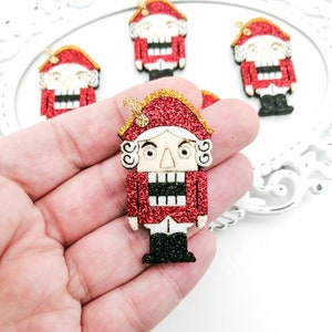 Peut inclure: Figurines de casse-noisette en paillettes rouges avec un chapeau en paillettes dorées et des bottes en paillettes noires. Le casse-noisette a un visage blanc avec des yeux noirs et une moustache noire.