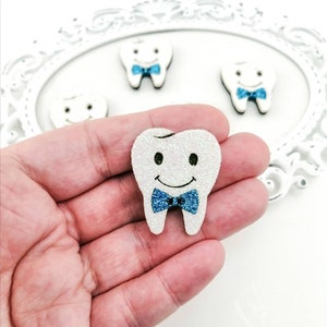 May include: A white glitter tooth with a blue bow tie. The tooth is smiling and has black eyes. The tooth is being held in a hand.