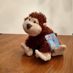 Puede incluir: Un mono de peluche marrón y beige peludo sentado en una superficie de madera. El mono tiene una etiqueta pegada en la espalda que dice "Webkinz"