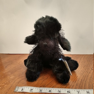 May include: Black plush poodle toy with a green tint. The toy is sitting on a wooden surface with a measuring tape in the foreground.