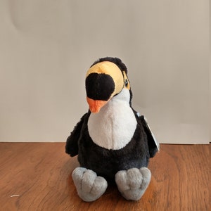 May include: A plush toucan bird toy with a black body, white chest, and grey feet. The toucan has a large orange and black beak and a yellow head. The toy is approximately 15 cm tall.